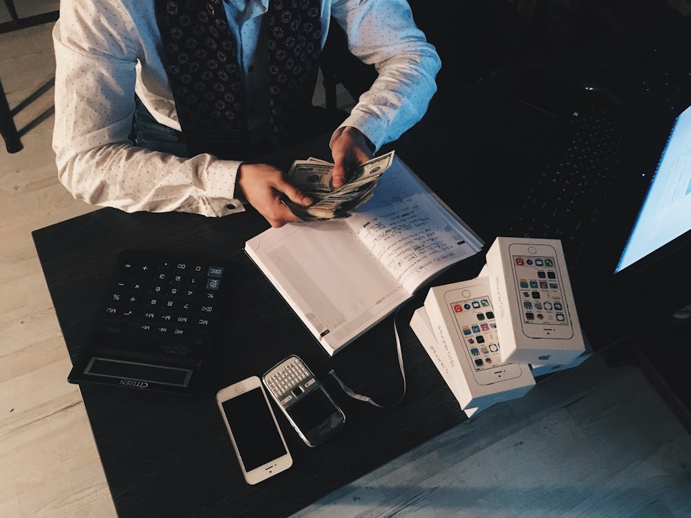 Business owner counting cash at a desk with a notebook, calculator, and smartphone boxes.