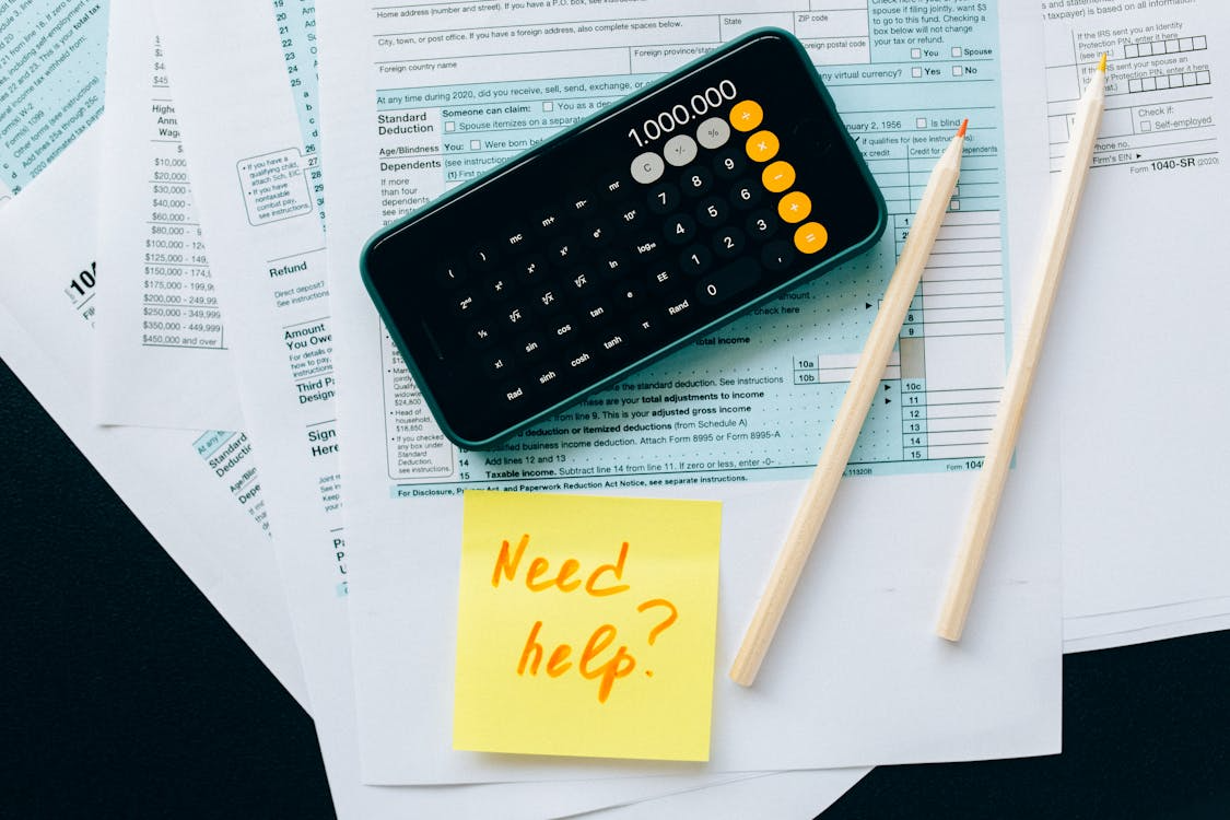 Smartphone with calculator app on tax documents beside pencils and sticky note reading “Need help?”.
