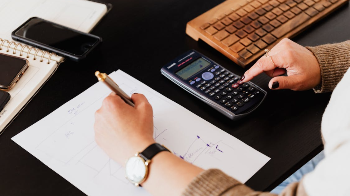 Picture13 Person calculating finances with a calculator and pen on a desk beside a notebook and smartphone.